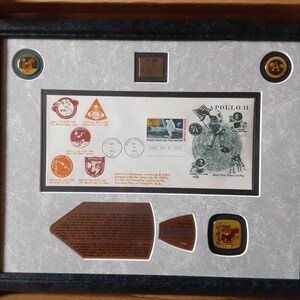 May include: A framed display featuring a commemorative Apollo 11 stamp, a small metal plaque, and several round, gold-colored medallions. The stamp depicts an astronaut on the moon with the text "First Man on the Moon" and "First Day of Issue". The plaque has a metallic finish and the medallions feature images of the Apollo 11 mission.