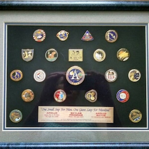 May include: A framed display of various NASA mission patches, including Apollo, Skylab, and Soyuz. The patches are arranged in a grid pattern on a black background. The frame is silver with a black mat and a blue outer frame. The text "One Small Step For Man, One Giant Leap For Mankind" is printed below the patches.