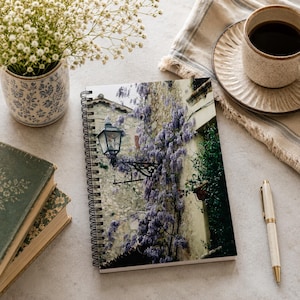 May include: A spiral-bound notebook featuring a photograph of a stone building covered in purple wisteria. A white and gold pen, a cup of coffee, a small vase of white flowers, and two antique books are also on the table.