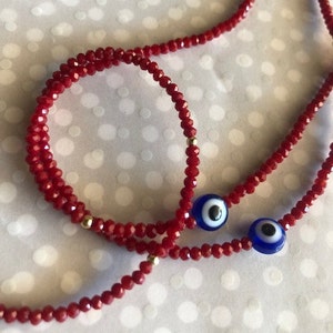 May include: A red beaded necklace and bracelet set with blue evil eye charms. The necklace and bracelet are made of small, red glass beads.