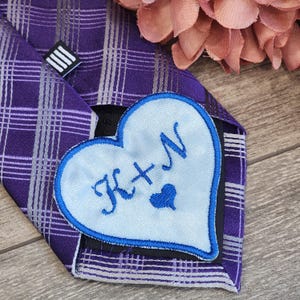 May include: A purple and silver plaid tie with a blue heart-shaped patch that says "K+N" in cursive with a small heart in the middle.