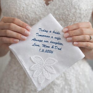 May include: White handkerchief with embroidered text in navy blue: "Today a bride, Tomorrow a wife, Always our daughter. Love, Mom & Dad 1.3.2026." Features a white floral lace design. Wedding keepsake.