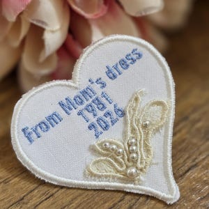 May include: White heart-shaped patch with blue embroidered text reading "From Mom's dress 1981 2026." The patch features a cream-colored lace design with pearl accents. The patch is on a wooden surface.