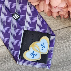 May include: A purple plaid tie with a light gray stripe pattern. The tie has a black label with two embroidered light blue hearts with the letters "K" and "N" in gold. The tie is made of a smooth fabric.