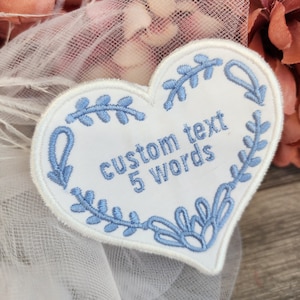 Puede incluir: Parche blanco en forma de corazón con borde floral bordado en azul y texto "custom text 5 words". Ideal para personalizar ropa o accesorios.