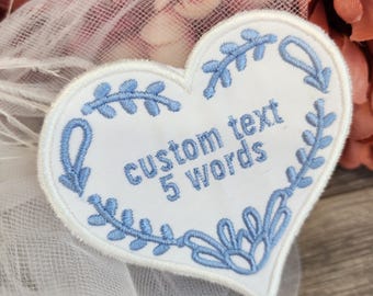 May include: A white heart-shaped patch with blue embroidered floral border and text that reads "custom text 5 words". The patch is likely for clothing or accessories.