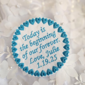 May include: White fabric circle with a blue embroidered border of hearts. The text "Today is the beginning of our forever. Love, Julie 1.19.25" is embroidered in blue on the circle.