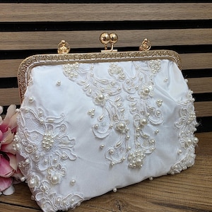 Peut inclure: Pochette en satin blanc ornée de dentelle et de perles. Le sac rectangulaire est doté d'un fermoir et d'un cadre dorés. La dentelle forme des motifs floraux et abstraits, avec des perles dispersées pour la texture.
