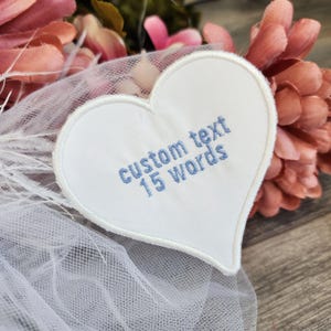 Könnte beinhalten: Weißer herzförmiger Stoff-Patch mit blau gesticktem Text, der "custom text 15 words" lautet. Der Patch befindet sich auf einem weißen Tüllstoff.