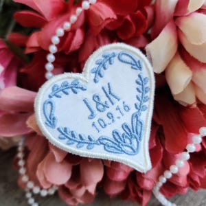 May include: A white heart-shaped patch with blue embroidery. The patch features a floral design and the text "J&K 10.9.16".