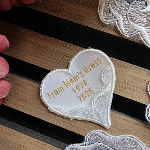 May include: A white heart-shaped patch with gold embroidered text reading "From Mom's dress 1974 2025". The patch has a delicate lace border. The background features a striped pattern and floral accents.