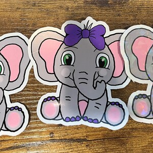 Puede incluir: Tres pegatinas de un elefante de dibujos animados con orejas rosas, un lazo morado y ojos verdes. El elefante está sentado con la trompa en la boca. Las pegatinas están sobre una superficie de madera.