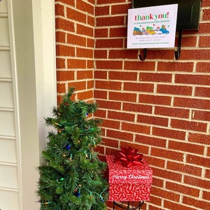 Puede incluir: Un peque&ntilde;o &aacute;rbol de Navidad con luces est&aacute; al lado de una caja de regalo roja con un lazo rojo y las palabras "Feliz Navidad" en ella. Un letrero en la pared de ladrillo dice "&iexcl;Gracias! A nuestros incre&iacute;bles repartidores! Apreciamos todo su trabajo duro y todo lo que hacen. Por favor, tomen algunas golosinas para el camino."