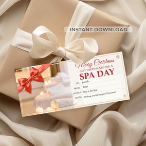 May include: A spa day gift certificate with a red ribbon, placed on a wrapped gift with a cream-colored bow. The certificate reads "Merry Christmas, Gift Certificate for a Spa Day". The gift is on a white fabric.