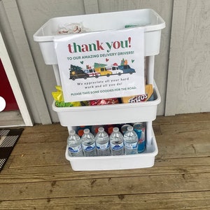 Puede incluir: Un carro de tres niveles blanco con ruedas con un cartel que dice "&iexcl;gracias! A nuestros incre&iacute;bles repartidores!" El carro est&aacute; lleno de bocadillos y bebidas, incluyendo botellas de agua, patatas fritas y dulces. El cartel tambi&eacute;n dice "&iexcl;Apreciamos todo tu trabajo duro y todo lo que haces! Por favor, toma algunas golosinas para el camino."
