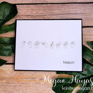 May include: A white card with black border features line drawings of hands spelling out a name in American Sign Language. The name "Madison!" is printed below the hand signs. The card is on a wooden surface with green leaves.