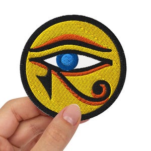 Könnte beinhalten: Ein gelber gestickter Aufnäher mit einem blauen Auge und orangefarbenen und schwarzen Details. Das Auge ist von einem schwarzen Kreis umgeben.