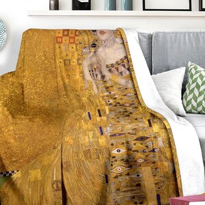 Puede incluir: Una manta de tiro dorada y marrón con una pintura de una mujer con un vestido dorado. La pintura es una reproducción de "El Beso" de Gustav Klimt.