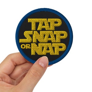 Op de afbeelding: Een ronde, zwarte patch met blauwe stiksels rond de rand. De tekst "TAP SNAP OR NAP" is geborduurd in geel.