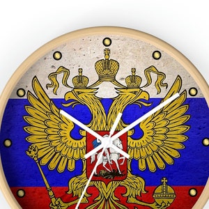 May include: A round wooden clock with a white face and black hands. The clock face features a stylized image of the Russian coat of arms, a double-headed eagle with a crown on its head, holding a scepter and orb. The eagle is set against a background of the Russian flag, with the colors red, blue, and white.
