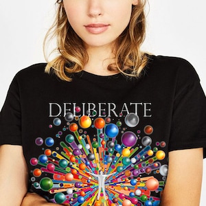 Puede incluir: Una camiseta negra con un gráfico colorido de una persona rodeada de orbes coloridos y la palabra "DELIBERATE" impresa en blanco.