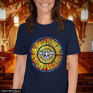 Könnte beinhalten: Ein marineblaues T-Shirt mit einem Buntglasdesign, das eine Taube in einem Kreis zeigt. Das Buntglasdesign ist in Gelb-, Orange-, Rot- und Blautönen gehalten.