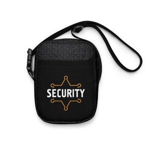 May include: Black crossbody bag with a textured top panel and a front pocket. The bag features the word "SECURITY" in white, inside a gold star-shaped design. It has a black adjustable strap and a side buckle.