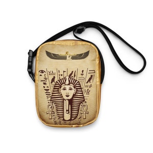 May include: A small brown and gold crossbody bag with an Egyptian pharaoh design. The bag features hieroglyphics, a winged sun disk, and an ankh symbol.