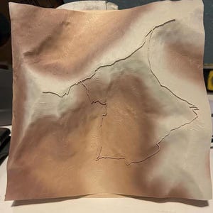May include: A textured, square-shaped piece of paper with a topographical map design. The paper has a gradient of colors, transitioning from light beige to a darker brown. The map's lines are raised, creating a 3D effect.