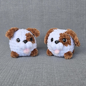 May include: Two round, plush, crocheted dog toys made of soft white and brown yarn. Each toy features large, glossy black eyes, a small black nose, and a tiny pink tongue protruding from the mouth. The dogs have floppy brown ears and paws.