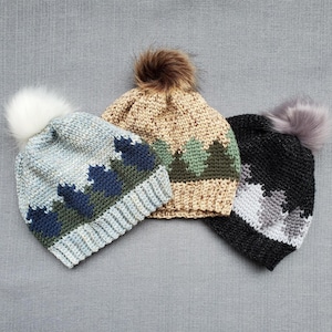 Könnte beinhalten: Drei gehäkelte Mützen mit Bommeln. Die Mützen sind in Blau, Beige und Schwarz mit einem geometrischen Muster.