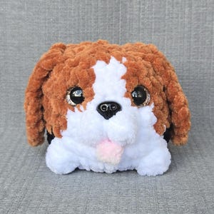 May include: A plush toy shaped like a dog, crafted from soft brown and white yarn. The stuffed animal features large, round, dark brown eyes with glittery accents, a black plastic nose, and a small, light pink tongue protruding from its mouth.