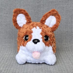 Könnte beinhalten: Ein Plüschtier in Form eines Hundes aus braunem und weißem Garn. Das Stofftier hat große schwarze Kunststoffaugen, eine schwarze Nase und eine kleine herausgestreckte rosa Zunge. Die Ohren sind aufrecht und spitz mit weißen Innenseiten.