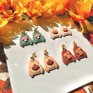 May include: A selection of handmade polymer clay earrings shaped like jumpers. The earrings showcase autumn-themed designs such as mushrooms, pumpkins, maple leaves, and squirrels. Each earring has a gold-coloured post.