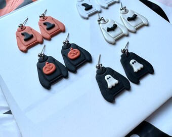 May include: A collection of Halloween-themed earrings. The earrings are shaped like sweaters, with designs including a witch hat, pumpkin, bat, and ghost. The sweaters are in various colors, including black, white, and orange.