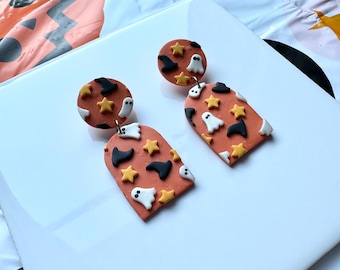 May include: A pair of Halloween-themed earrings. The earrings feature a burnt orange base with white ghost, black witch hat, and yellow star designs. The earrings are on a white surface.