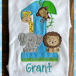 May include: A white onesie with a blue number one, a green leaf, a yellow giraffe, a brown monkey, a gray elephant, a brown lion, and the name "Grant" embroidered on it.