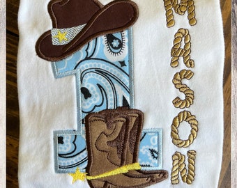 May include: A white onesie with a blue and white paisley pattern number one, a brown cowboy hat, and brown cowboy boots. The name "Mason" is embroidered in gold rope on the right side of the onesie.