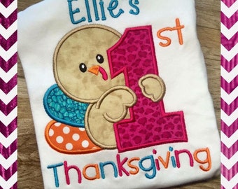 May include: A white onesie with a brown turkey holding a pink number one with a leopard print design. The onesie also has the text "Ellie's 1st Thanksgiving" embroidered on it.