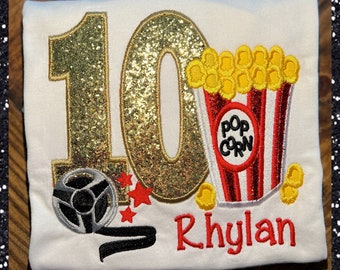 May include: A white t-shirt with a gold glitter number 10, a red and white popcorn bucket, a silver film reel, and red stars. The name "Rhylan" is embroidered in red below the design.