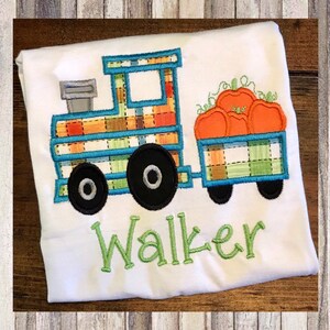 May include: A white t-shirt with a blue and green plaid train pulling a wagon full of orange pumpkins. The wagon is also plaid. The name "Walker" is embroidered in green below the train.