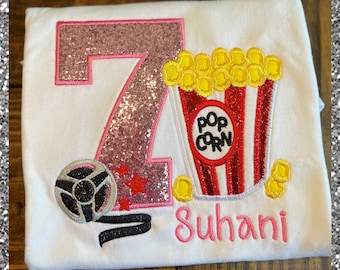 May include: Pink glitter number seven with a red and white popcorn bucket and a black film reel. The name "Suhani" is embroidered in pink below the popcorn bucket.