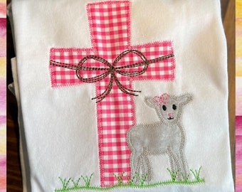 May include: A white t-shirt with an embroidered pink and white gingham cross with a brown bow and a gray lamb standing in front of it. The lamb has a pink flower on its head.