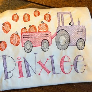 May include: A white t-shirt with an embroidered design of a purple tractor pulling a red wagon full of orange pumpkins. The pumpkins are scattered around the tractor. The name "Pinklee" is embroidered below the tractor.