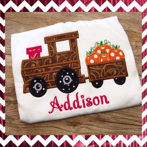 May include: A white t-shirt with an embroidered brown train pulling a pumpkin-filled wagon. The train has a pink box on the front and the name "Addison" is embroidered in pink below the train.