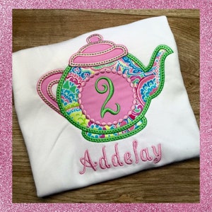May include: A white t-shirt with a pink and green embroidered teapot design. The teapot has a pink number "2" in the center. The name "Addalyn" is embroidered below the teapot.