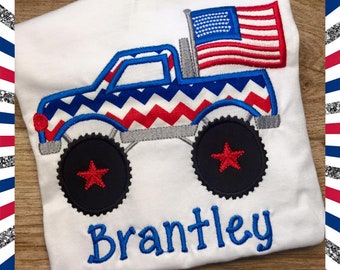 May include: White t-shirt featuring an embroidered monster truck design with patriotic red, white, and blue colors. The truck has large black wheels with red star accents and an American flag. The name "Brantley" is embroidered in blue.