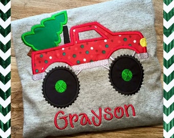 May include: A gray t-shirt featuring a red truck with a Christmas tree in the back. The truck has black wheels with green centers and is decorated with white polka dots. The name "Grayson" is embroidered in red.