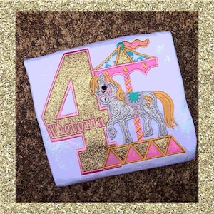 May include: A white t-shirt with a gold glitter number "4" and the name "Victoria" embroidered on it. A pink and white carousel horse is embroidered next to the number.