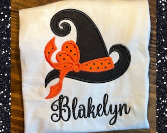 May include: White t-shirt featuring a black witch hat with an orange bow and polka dot ribbon. The name "Blakelyn" is embroidered in black script below the hat. A festive Halloween-themed design.